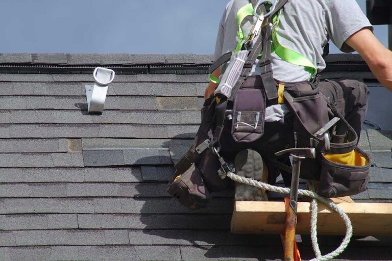 Roof inspector examining curled shingles and flashing on a residential roof beside a snow-covered yard.